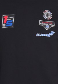 Camiseta negra con parches bordados: "1 CLSA LDN", "CLOSURE RACING TEAM 15" y un logo de globo en colores azul, rojo y blanco.
