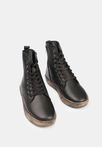 Botas de cuero negras hasta el tobillo con puntera redonda, parte delantera con cordones y cremallera lateral. Suela de goma texturizada con un borde marrón claro en contraste.