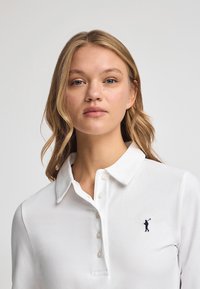 Vit pikétröja i bomull med klassisk krage, knapplist och en broderad logotyp av en figur som håller en golfklubba på bröstet.