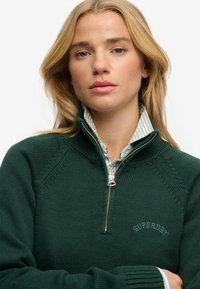 Dunkelgrüner, wärmender Strickpullover mit Reißverschluss und Rippstruktur, ausgestattet mit einem hohen Kragen und gesticktem "SUPERDRY"-Logo in Hellblau. Gestreiftes Hemd darunter.
