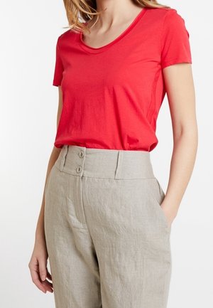 Woman wearing a red short-sleeve top tucked into high-waisted beige linen trousers with a double-button waistband.