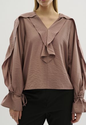 Femme portant un chemisier mauve avec col en V volanté et manches bouffantes nouées aux poignets, associé à un pantalon noir, sur fond clair.