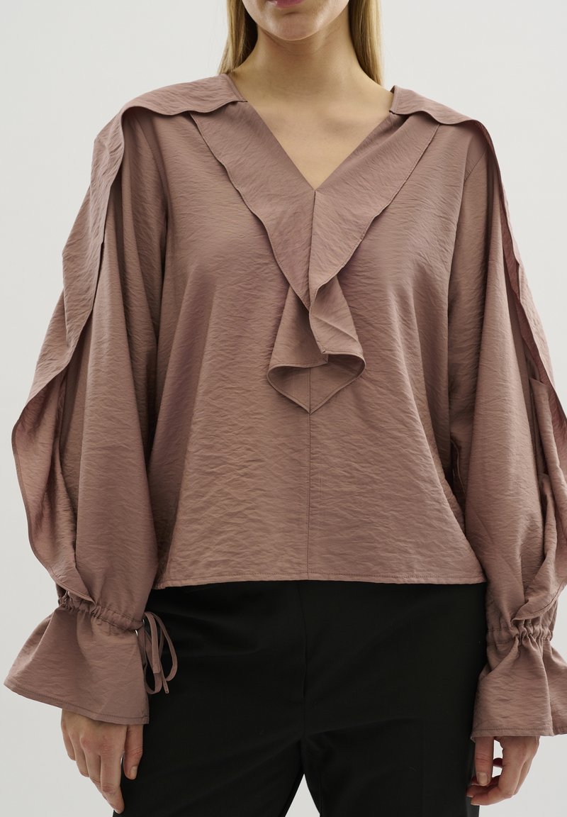 Femme portant un chemisier mauve avec col en V volanté et manches bouffantes nouées aux poignets, associé à un pantalon noir, sur fond clair.