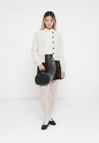 Jonge vrouw in een witte cardigan, zwarte leren rok, witte maillot en zwarte ballerina's, met een kleine zwarte handtas tegen een witte muur.