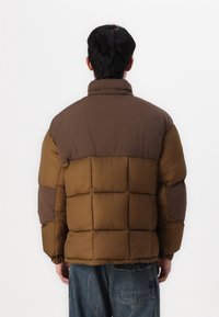 Braune Pufferjacke mit einem kontrastierenden dunkelbraunen oberen Bereich, der ein gestepptes Design, elastische Bündchen und einen hohen Kragen aufweist.