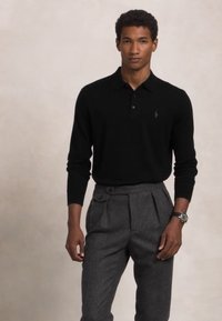 Črna polo majica z dolgimi rokavi in tri gumbi, v kombinaciji s sivo plisirano hlačami in ure, pozira na nevtralnem ozadju.