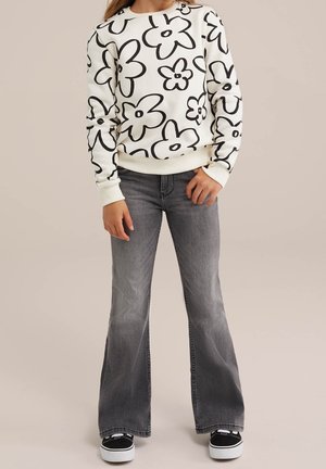 Enfant portant un sweat-shirt blanc avec une grande impression de fleur noire, un jean évasé gris et des baskets noires à semelles blanches.