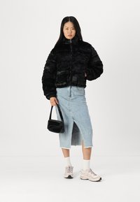 Jachetă din blană sintetică neagră cu fermoar, asortată cu o fustă din denim bleu deschis, o geantă neagră și teniși roz. Fundal simplu.