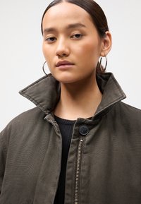 Young woman with slicked-back hair wears large silver hoop earrings and an olive green jacket with a popped collar and visible zipper.