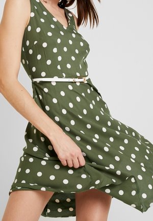 Grøn ærmeløs kjole med hvide polkaprikker. Har V-hals, tætsiddende talje og et hvidt bælte med guldspænde. Glat stof.