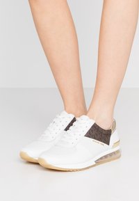 Sneaker bianco con una tomaia in mesh traspirante, dettagli marroni a motivo, hardware dorato e suola con cuscino d'aria trasparente.