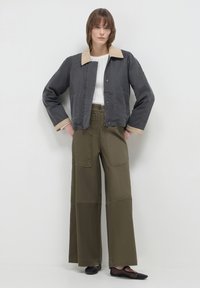 Donna in piedi contro un muro bianco, indossa un giacchetto grigio scuro con colletto beige, un top bianco a maglia, pantaloni larghi oliva e ballerine nere.