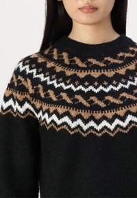 Pull en maille noir avec un motif décoratif sur le yoke en marron, blanc et beige, présentant des motifs en zigzag et géométriques.
