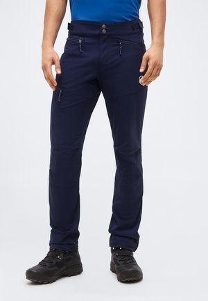 Άνδρας που φοράει σκουρόχρωμο μπλε παντελόνι πεζοπορίας με τσέπες με φερμουάρ και μαύρα παπούτσια πεζοπορίας, στέκεται μπροστά σε ένα απλό φόντο.