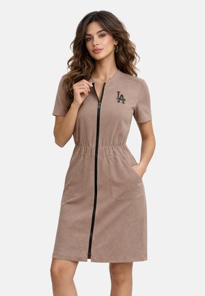 Kobieta z długimi falowanymi włosami w brązowej sukience o krótkich rękawach sięgającej kolan, z zamkiem z przodu i logo LA na piersi, ręką w kieszeni.