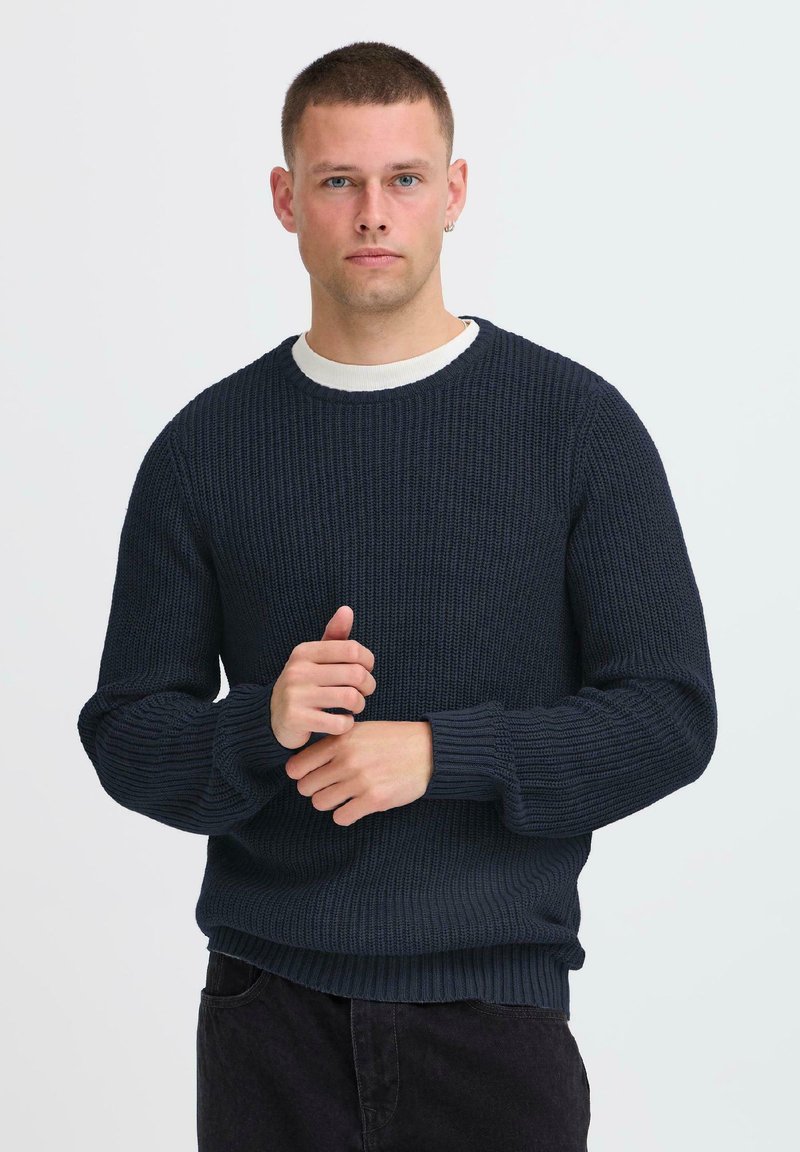 Young man with short hair wearing a navy blue knitted sweater and black pants, standing against a plain light background.