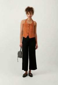 Πορτοκαλί σουέτ γιλέκο με εφαρμοστό σχέδιο, κουμπώνοντας μπροστά, συνδυασμένο με φαρδιά μαύρα παντελόνια και μία υφασμάτινη μαύρη-άσπρη τσάντα.