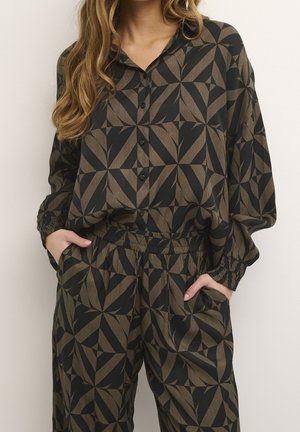 Vrouw draagt loszittend zwart en bruin geometrisch patroon blouse met knopen en bijpassende broek met elastische taille, handen in de zakken.