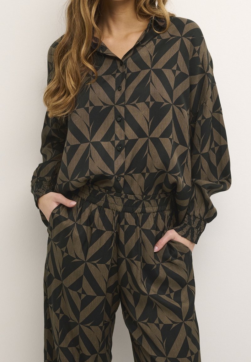 Femme portant une chemise ample noire et marron à motifs géométriques avec boutons, et un pantalon assorti à taille élastique, mains dans les poches.