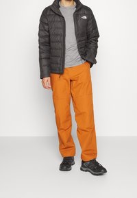 Svart pufferjacka, grå t-shirt och orange byxor. Svarta sneakers med grå detaljer. Stående framför en neutral bakgrund.