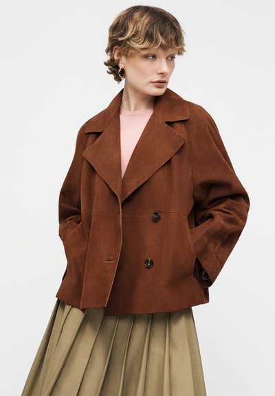 Γυναίκα με κοντά κυματιστά μαλλιά που φοράει μεγάλα κρίκους σκουλαρίκια, καφέ σουέντ διπλόκουμπο σακάκι, ροζ τοπ και μπεζ πλισέ φούστα, με τα χέρια στις τσέπες.