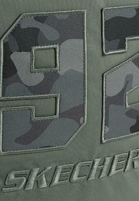 Tela verde con grandes números bordados "92" en un patrón de camuflaje con hilos texturizados, junto al nombre de la marca "SKECHERS" en letras gruesas y bordadas.