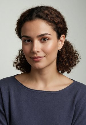 Kvinde iført en marineblå sweater med bred udskæring; krøllet hår; en lille kors-ørering med perleaccent. Glat hud og neutral baggrund.
