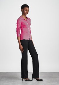 Cardigan en tricot magenta avec des manches longues et des boutons, associé à un pantalon noir à jambes droites et des escarpins noirs à bout pointu sur un fond neutre.