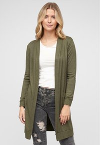 Oliivinvihreä neulecardigan, jossa on avoin etuosa, pitkät hihat, ribbireunukset ja sivutaskut, yhdistetty valkoisen paidan ja kulutettujen tummien farkkujen kanssa.