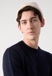 Mornarsko pleten pulover z rebrastim ovratnikom in šivi na ramenih. Model nosi svetlo bež rebrasto kapo. Jasna ozadje izboljša osredotočenost na oblačila.