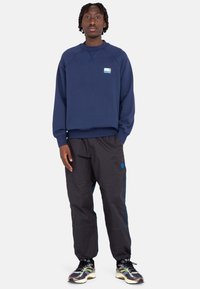 Sudadera azul marino con un pequeño logo, combinada con pantalones negros que tienen bolsillos laterales y acentos en azul. Llevado con zapatillas negras y verdes.