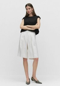 Czarny top z krótkim rękawem w połączeniu z białymi plisowanymi szortami o wysokim stanie. Model nosi wzorzyste baleriny w szpic. Prosty, nowoczesny design.