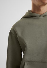 Felpa con cappuccio verde oliva, realizzata in tessuto morbido e strutturato. Presenta una tasca a marsupio e una vestibilità comoda con maniche raglan.