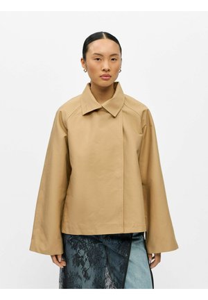 Kvinde med glat sort hår iført en beige jakke med brede ærmer og en blå nederdel med sort blonder over et hvidt baggrund.