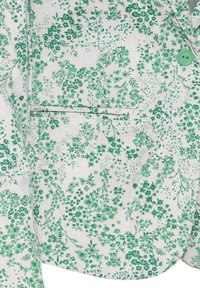 Tissu blanc avec motif floral vert sur une veste montrant un bouton vert et une poche fendue horizontale.