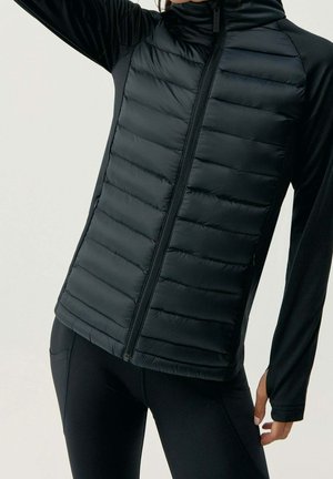 Henkilö pukeutuneena mustaan, vetoketjulliseen tikattuun takkiin, jossa on pitkät hihat, ja mustiin tiukasti istuviin leggingsseihin, yksinkertaisen taustan edessä.