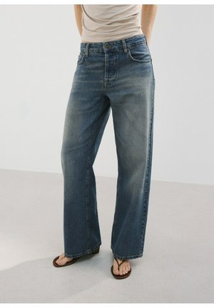 Persona che indossa jeans larghi blu scoloriti con bottone e cerniera frontale, abbinati a infradito scuri, in piedi su un pavimento chiaro.