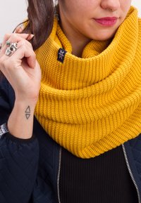 Knit, yellow cowl scarf features a textured ribbed design with a tag accent. Worn with a black top and blue padded jacket.