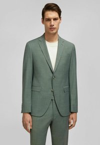 Lichtgroen, op maat gemaakt colbert van een gestructureerde stof, met twee voorkanten zakken, notch-revers en twee knopen. Gecombineerd met een effen overhemd.