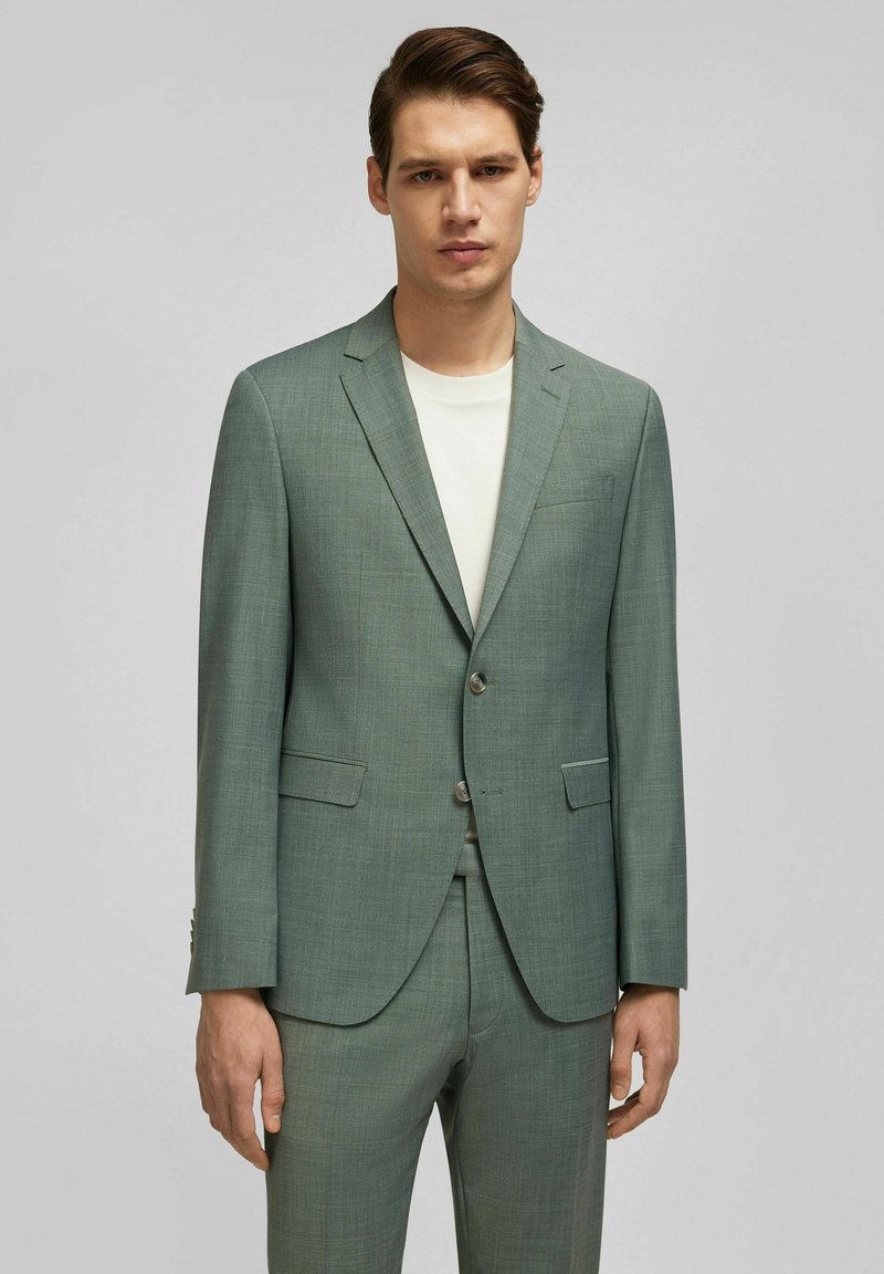 Lichtgroen, op maat gemaakt colbert van een gestructureerde stof, met twee voorkanten zakken, notch-revers en twee knopen. Gecombineerd met een effen overhemd.