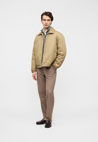 Veste matelassée beige avec une couche intérieure grise, col à boutons, portée par-dessus une chemise claire et un pantalon marron, associée à des chaussures marron foncé.
