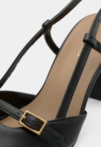 Chaussure à talon haut noire en cuir, avec des brides croisées fines, une boucle dorée et une semelle intérieure beige, mettant en avant un design minimaliste.