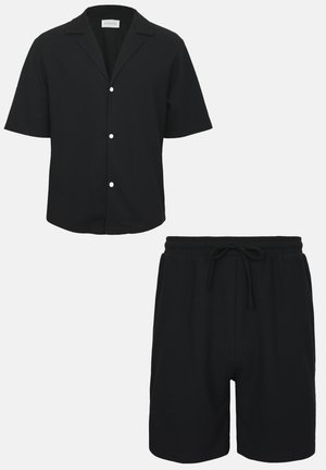 Chemise noire à manches courtes avec boutons blancs associée à un short noir à cordon sur un fond uni.