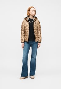 Νεαρή γυναίκα στέκεται φορώντας μπεζ καπιτονέ μπουφάν, μαύρο πουκάμισο, κολιέ με πέρλες, μπλε τζιν και μπεζ παπούτσια, μπροστά σε λευκό φόντο.