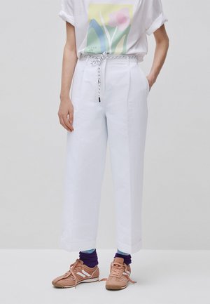 Persona con pantalones blancos cortos de cintura alta con un cinturón de cuerda, una camiseta blanca con estampado, calcetines morados y zapatillas marrón claro.