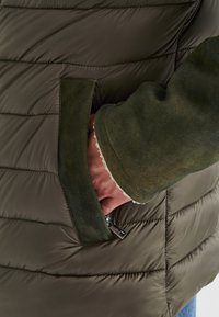 Hand in zipper pocket einer olivgrünen steppjacke mit Wildlederbesatz an den Bündchen und Taschen, die weiche Stoffstruktur zeigt.