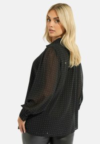 Blusa nera trasparente con maniche lunghe e gonfie, decorata con pois in foglia d'argento. Presenta un colletto e polsini con bottoni.