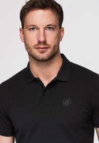 Homme aux cheveux bruns courts et barbe, portant un polo noir avec un petit logo brodé sur la poitrine.