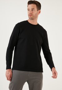 Fekete, hosszú ujjú, kerek nyakú póló, texturált anyagból készült, bordás mandzsettákkal és aljjal. Szürke nadrággal viselve, amely lazább szabású.