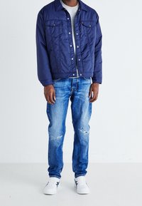 Mørkeblå quiltet jakke med knaplukning og to forlommer, kombineret med slidte lyseblå jeans og hvide sneakers.